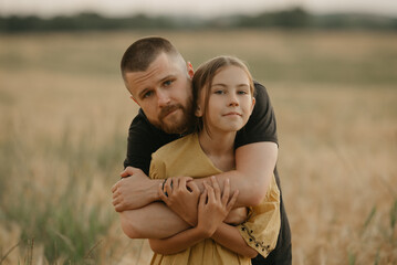 A father with a beard is hugging the shoulders of his daughter in the center of the wheat field at sunset. Happy family of the farmer.