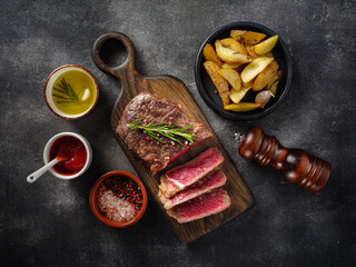 Sliced grilled meat steak New York Striploin with sauce and potato on wooden board on grey background. Top view.