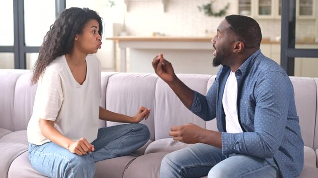 An African-American couple is quarreling sitting on the couch at home, misunderstands each other, irritated woman and man screaming to each other, arguing emotionally. Crisis and relationship problems