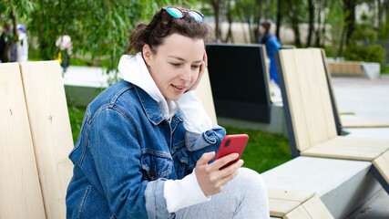 Pretty young woman using smartphone, looking at screen sitting outdoors on bench in city park.Checking social media, chatting with friend, reading news,watching online webinar using wireless internet