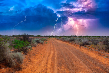 Naklejka na ściany i meble Deserted Australian outback landscape with red dirt road towards horizon with bushes in roadsides and heavy thunderstorm with white purple lightnings on the horizon