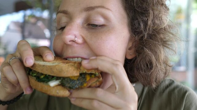 Young caucasian food-loving woman takes a healthy organic vegan sandwich, bites it with appetite and looks at it with appreciation 