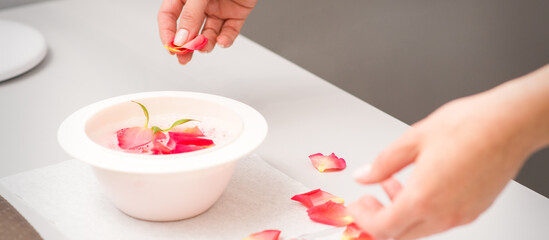 Fototapeta na wymiar Beautician's female hands preparing manicure bath with red and pink roses petals on the table in spa