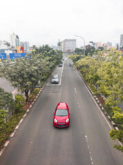 A beautiful red car crosses the middle of Jakarta Indonesia's road, on the left and right of the road, there are shady trees, making the road more comfortable for vehicles to pass.