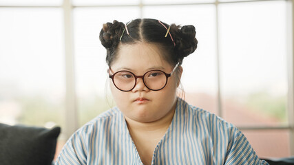 Pretty Down syndrome girl feels bored everything around her, sitting alone on the sofa with sad...