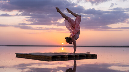 Beautiful graceful girl performs a handstand in the water of a salt lake. incredible pink-purple sunset in the mirror surface of the lake. sporty woman in dress