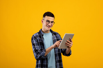 Online life concept. Funky young man using tablet pc on orange studio background