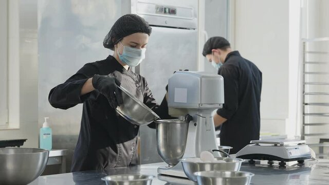 Concentrated young woman in Covid face mask pouring ingredients in automatic mixer with blurred man working on commercial kitchen at background. Workers in candy store on coronavirus pandemic.