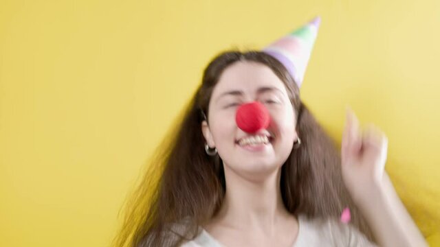 Portrait of a cheerful woman in a paper cap and a false red nose, who happily throws confetti and claps her hands. Yellow background. April's fool day concept