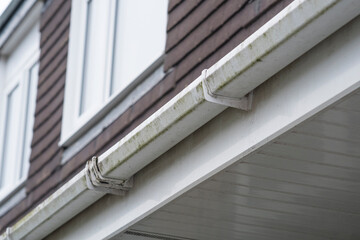 Dirty clogged white plastic pvc gutters and drain pipes with mossy green mould on plastic fascias.  Blocked drains and guttering need window cleaners and regular yard  maintenance for good drainage.