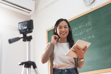 Young Asian woman teacher using digital camera standing online teaching with headphone and digital...