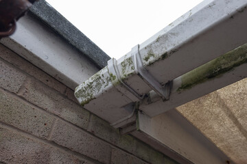 Dirty clogged white plastic pvc gutters and drain pipes with mossy green mould on plastic fascias. ...