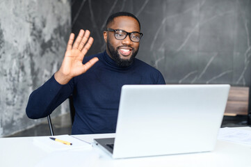 Positive African-American male employee using computer app on the laptop for video call sitting at...