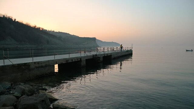 San Simon bay, Slovenia: father and son catching fish on concrete pier. Calm Adriatic Sea at sunset in winter. Small fishing boat on calm water. Static, real time, wide angle