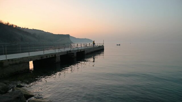 San Simon bay, Slovenia: father and son catching fish on concrete pier. Calm Adriatic Sea at sunset in winter. Small fishing boat on calm water. Static shot, real time, wide angle