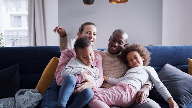Multi ethnic family with two small children on sofa at home, looking at camera.
