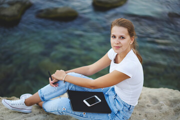 Teenage hipster girl with digital mobile device and education textbook, portrait of Caucaisan millennial woman dressed in trendy jeans looking at camera during rest chilling at seashore rock
