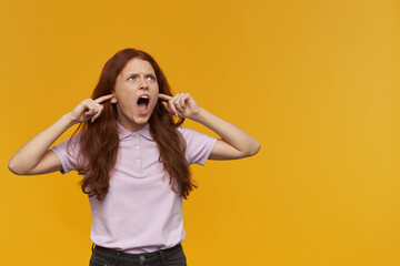 Fototapeta premium Angry, displeased woman with long ginger hair. Wearing pink t-shirt. Close ears with fingers and shouts, irritated with noise. Watching to the right at copy space, isolated over orange background