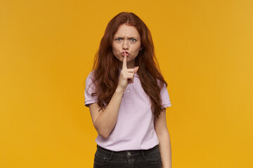 Unhappy girl, frowning redhead woman with long hair. Wearing pink t-shirt. Emotion concept. Showing silence sign, demand to be quiet. Watching at the camera, isolated over orange background
