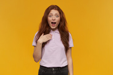 Cute, positive woman with long ginger hair. Wearing pink t-shirt. People and emotion concept. Pointing at herself. Introducing herself. Watching at the camera, isolated over orange background