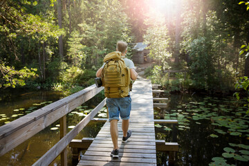 Mature man exploring Finnish nature in summer, walking across the bridge. Hiker with big backpack traveling in forests. Summer Scandinavian landscape of lakes and woods. 