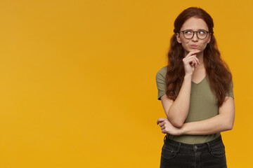 Portrait of thinking, dreaming lady with long ginger hair. Wearing green t-shirt and eyewear. Emotion concept. Touching her chin and watching to the left at copy space, isolated over orange background