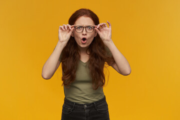 Surprised looking girl, shocked redhead woman with long hair. Wearing green t-shirt and glasses. Emotion concept. Touching her glasses. Watching at the camera, isolated over orange background