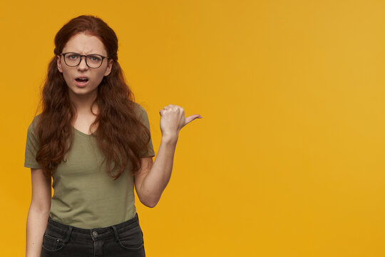 Confused girl, frowning redhead woman with long hair. Wearing green t-shirt and glasses. Watching at the camera and pointing with thumb to the right at copy space, isolated over orange background