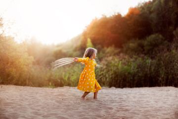 Caucasian cute little girl in yellow dress dancing on the sand in summer at sunset, happy childhood and freedom