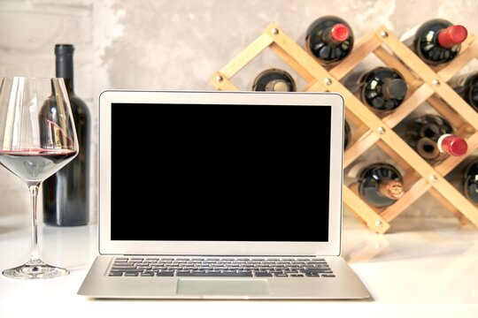 Empty Blank Screen Laptop Computer On Table With Wine Bottle Wine Rack Holder In The Background. Buying Wines Online, Home Delivery Concepts.