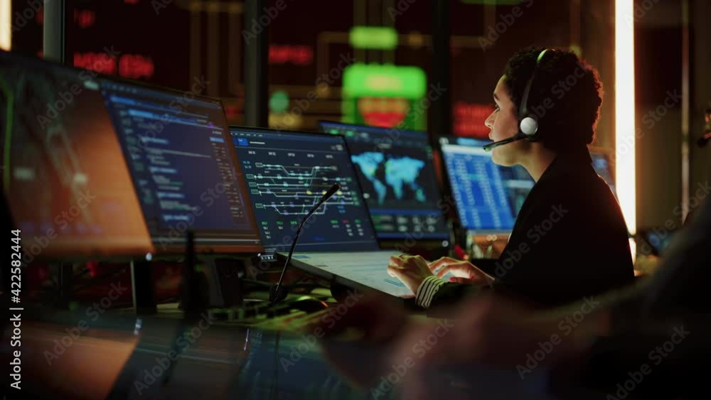 Poster Multiethnic Female IT Technical Support Specialist Talking with a Client in Headphones and Working on Desktop Computer in Monitoring Control Room with Big Digital Screens with Server Blockchain Data.