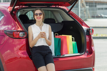 Smiling  young woman standing near red car with coffee and colourful paper bags. 