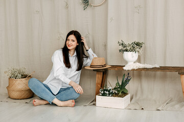 Studio photo of a girl wearing a shirt and blue jeans with bare feet. She rests on the bench, propping her head on her hand.Beige linen background wicker pots with white flowers