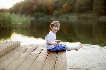 Caucasian cute boy on the footbridge by the lake, happy childhood in the village, child play in the park near the water, swing their feet in the water of the lake