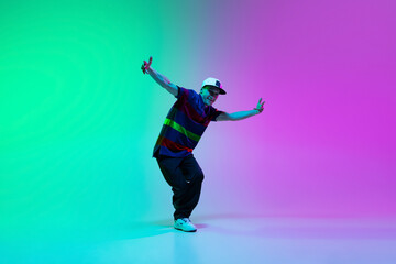 Fototapeta na wymiar Beautiful sportive boy dancing hip-hop in stylish clothes on colorful gradient background at dance hall in neon light.