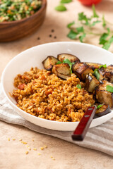 Bulgur pilaf (pilavi) with tomatoes and fried eggplants in a white plate on a light table close-up. Turkish cuisine, vegan food.