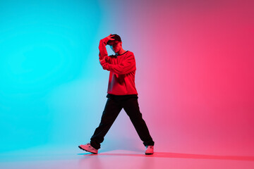 Beautiful sportive boy dancing hip-hop in stylish clothes on colorful gradient background at dance hall in neon light.