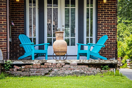 Two turquoise adirondack chairs and a chiminea on a crumbling brick porch in font of french doors on summer day with tiny American flag displayed