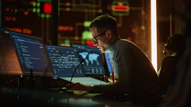 Professional IT Technical Support Specialist Making an Intercom Announcement on a Microphone and Working on Desktop Computer in Monitoring Control Room with Big Digital Screens with Server Data.