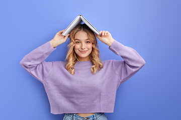 Joyful teen girl have fun holding book over head, looking at camera