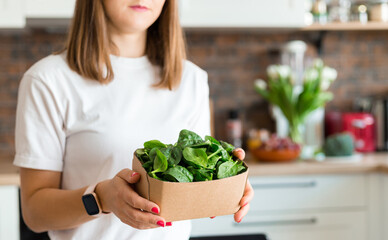 Woman in white t-shirt holding a green spinach in recycling ecology packing in kitchen at home. Raw food diet and healthy eating concept. Baby food