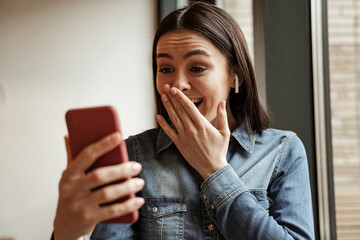 amazed woman in wireless earphones listening music and covering mouth while holding blurred smartphone