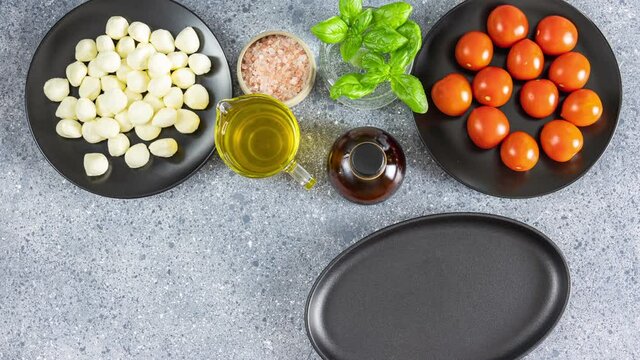 Making of Caprese salad with cherry fresh tomatoes, mozzarella cheese and basil served on oval black plate on light gray table surface, top view. Traditional Italian food. Stop motion animation