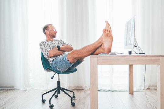 A middle-aged man in eyeglasses watching a modern computer in his living room. He put her barefoot lags on the table and relaxing. Video streaming on worldwide quarantine time concept