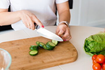 Brunette Woman in white t-shirt preparing vegetable salad in the kitchen at home, Wife Cutting cucumber on table. Healthy Food, Vegan Salad. Dieting Concept.