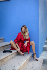 Fabulous female in red dress sitting at the staircase next to painted blue wall