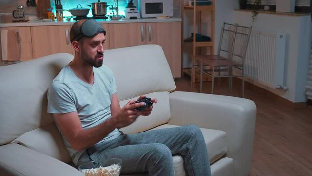 Frustrated Man With Sleep Mask Losing Videogame Competition Using Wireless Controller. Disappointed Pro Gamer Lying On Sofa In Front Of Television Late At Night In Kitchen