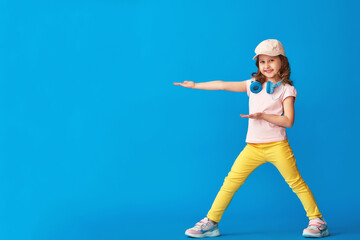 Fototapeta na wymiar little girl in summer clothes and headphones poses on blue background in full growth and looks to side and points her hand to side. concept summer and spring casual children's clothing. Copy space