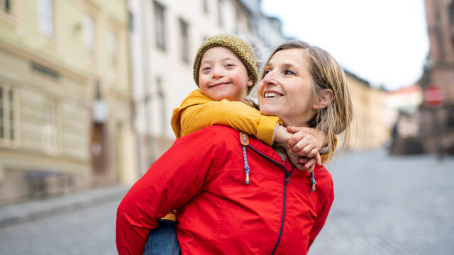 Single Mother Giving Piggyback Ride To Down Syndrome Child Outdoors In Town.