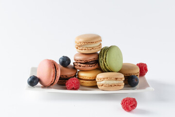 Multi colored french macaroons and berries such as raspberry and blueberry on a plate over white background.